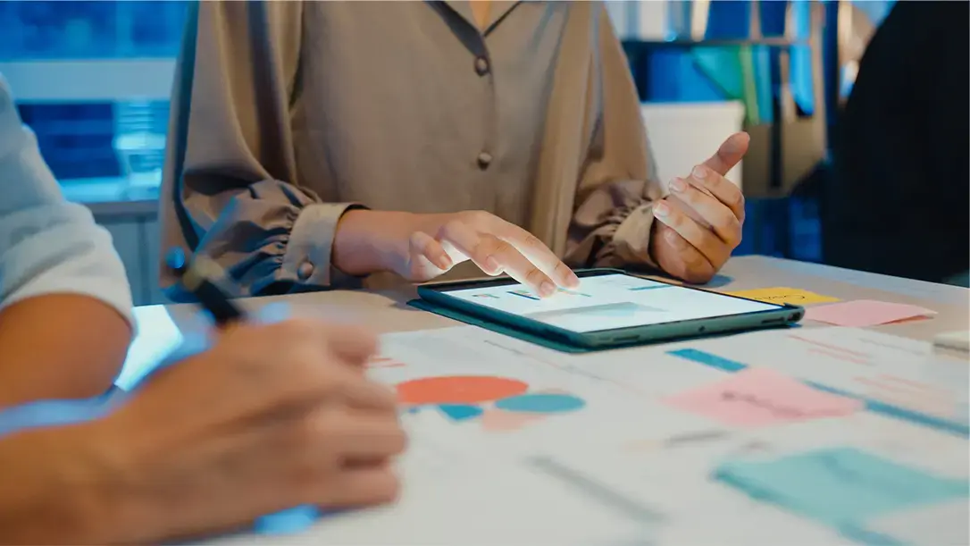 Persona utilizando una tablet táctil sobre una mesa con gráficos y notas adhesivas durante una reunión de trabajo